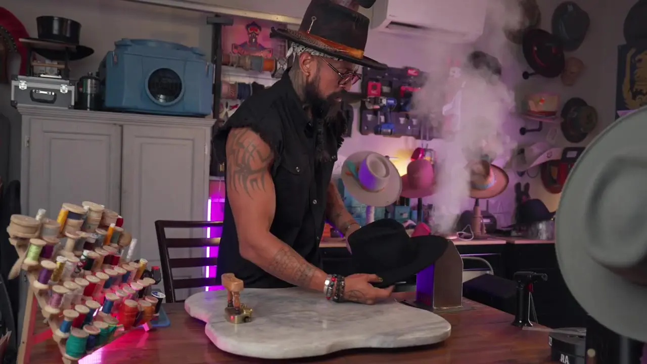 La Sombra shapes a black fur felt hat beside an active workshop steamer as steam rises.